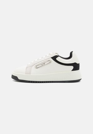 White sneakers with black and grey accents, leather and suede materials, lace-up design, cushioned sole, and a distinctive side zipper detail.