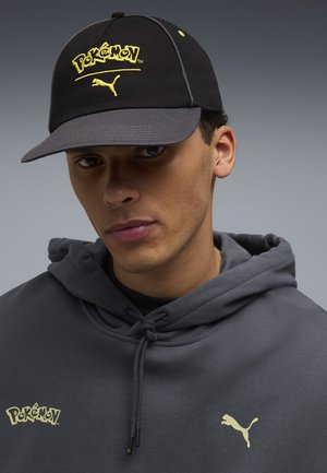 Jeune homme portant une casquette noire et grise avec les logos jaunes Pokémon et Puma, et un sweat à capuche gris assorti avec les logos Pokémon et Puma.