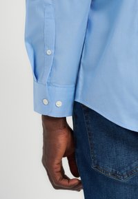 Σκούρο δέρμα χεριού που ακουμπάει στο πλευρό, φοράει ανοιχτό μπλε πουκάμισο με κουμπωτές μανσέτες και σκούρο μπλε τζιν τζιν με ορατές ραφές στην πίσω τσέπη.