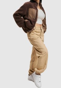 Giacca in pile marrone con tasca con zip, pantaloni cargo beige e sneaker bianche. La giacca ha un colletto alto; i pantaloni hanno polsini elasticizzati e una vestibilità rilassata.
