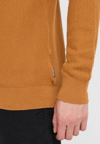 Pomarańczowy sweter dzianinowy z teksturowanym wzorem, z ściągaczami przy mankietach i u dołu, oraz małą metką marki z boku. Widać czarne spodnie.