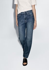 Mörkblå rakbenade jeans med midjehöjd, fem fickor, som bärs med en vit skjorta och svarta lädersandaler.