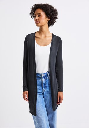 Cardigan noir à manches longues avec un devant ouvert, en tissu léger, associé à un haut blanc et un jean taille haute bleu, sur fond uni.