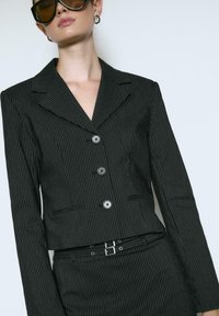 Sort stribede blazer med klapkrave, tre-knaps lukning og forlommer. Parret med en matchende nederdel og bælte.