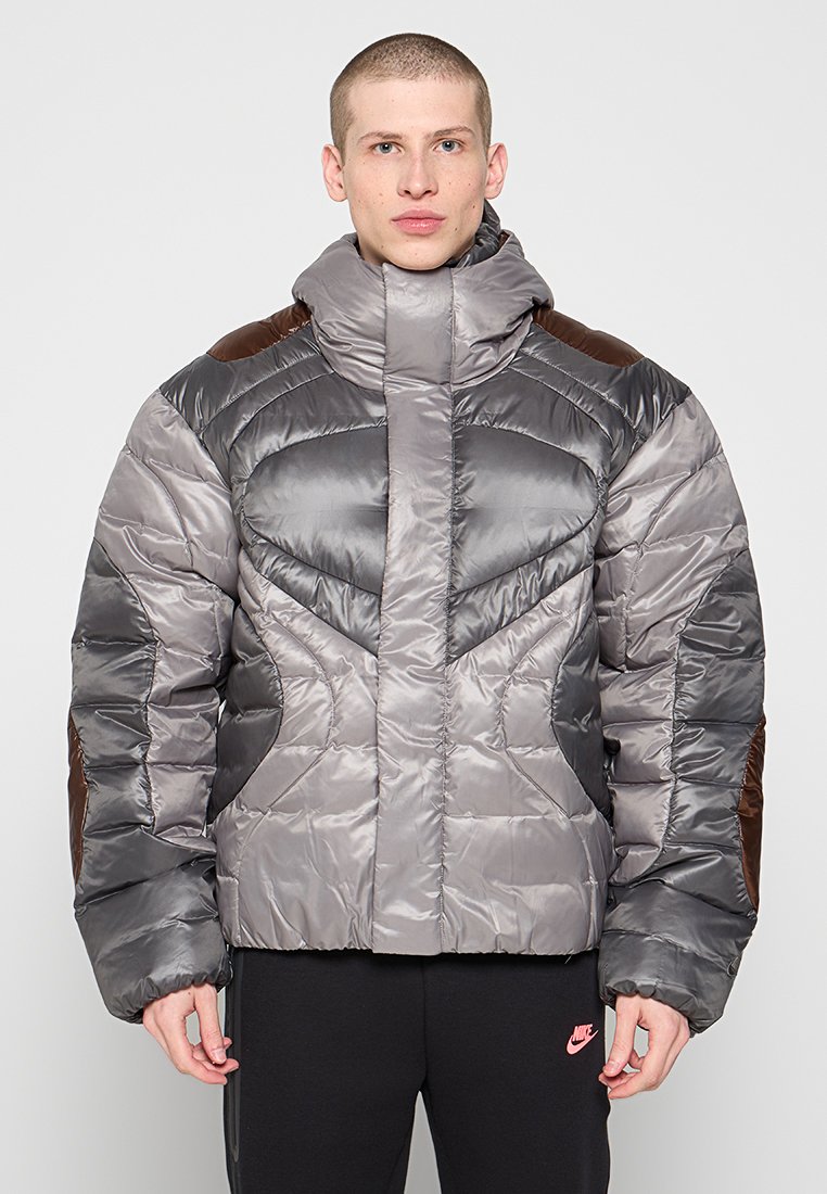 Chaqueta acolchada en tonos de gris con acentos marrones, que presenta un cuello alto, textura acolchada y una silueta cuadrada.