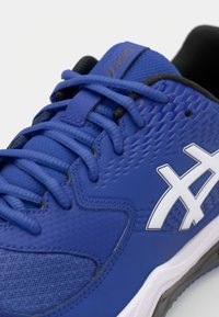 Chaussure de sport bleue avec tige en caoutchouc texturé, logo blanc contrastant, lacets plats et col de cheville rembourré. Design ajusté pour un soutien optimal.