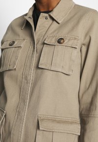 Chaqueta de lona color beige con dos bolsillos en el pecho, botones a presión y cremallera frontal. Presenta un cuello y un corte relajado. Textura de tela duradera.