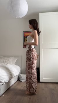 Γυναίκα όρθια πλάγια σε υπνοδωμάτιο φορώντας φλοράλ παντελόνια και λευκό τοπ χωρίς τιράντες, με λευκό κρεβάτι, ντουλάπα και αφίσα στο παρασκήνιο.