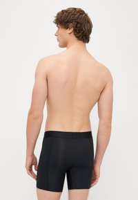 Μαύρα μποξεράκια με υφασμάτινη ζώνη που εμφανίζει το όνομα της μάρκας. Λείος ύφασμα, σφιχτή εφαρμογή, μήκος μέχρι το μισό μηρό, και χωρίς επιπλέον σχέδια.