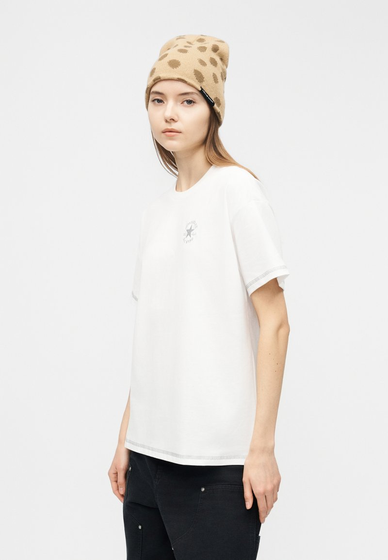 Giovane che indossa un berretto beige con stampa leopardata, una maglietta bianca larga con un piccolo logo e pantaloni neri, in piedi davanti a uno sfondo bianco uniforme.