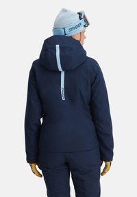 Marineblaue Skijacke mit Kapuze und hellblauen Akzenten, die einen vertikalen Streifen auf dem Rücken und ein schlankes, enges Design aufweist. Goldene Handschuhe sichtbar.