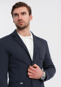 Man med kort brunt hår som bär en marinblå kavaj över en vit skjorta med krage och justerar kavajen med sin högra hand.