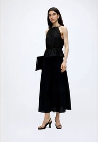 Γυναίκα με μαύρο αμάνικο halter top και μακριά φούστα, κρατώντας μαύρο φάκελο, στέκεται με μαύρα πέδιλα με τακούνια.