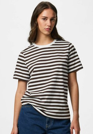 Ung kvinde iført en hvid og brun vandret stribet kortærmet skjorte og blå jeans, stående mod en ensfarvet baggrund.