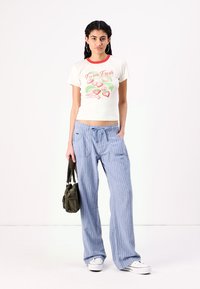 Calças largas listradas em azul com bolsos, combinadas com uma t-shirt creme com um gráfico de morango e bordas vermelhas. Bolsa de tecido verde.