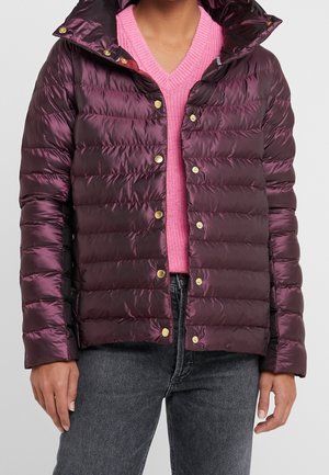 Personne portant une doudoune brillante violet foncé avec des boutons dorés sur un pull rose en maille à col en V et un jean gris foncé.