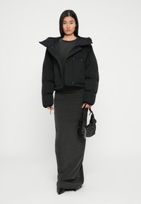 Zwarte pufferjas met een cropped ontwerp, ritsluiting en twee voorzakken; gecombineerd met een lange, aansluitende grijze jurk en een kleine tas met studs.