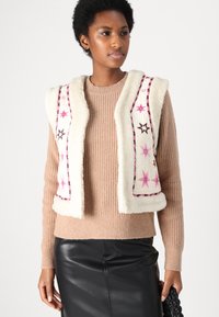 White faux fur vest with embroidered pink stars and black accents, layered over a light brown knitted sweater, paired with a black skirt.