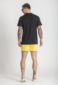 Černé tričko s krátkým rukávem, žluté plavkové šortky, lehký materiál, volný střih. Viditelné tetování na nohách. Model má na sobě sluneční brýle a nazouvací boty.
