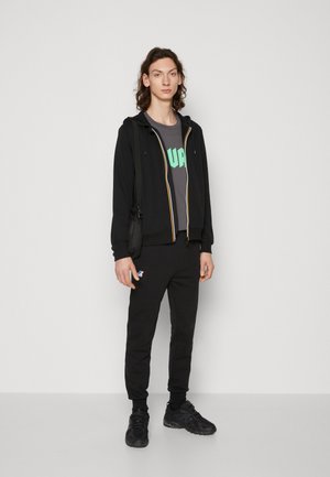 Felpa nera con zip e dettagli gialli sopra una t-shirt grigia con testo verde, abbinata a pantaloni jogger neri e sneaker nere.