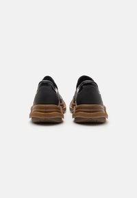 Une paire de baskets basses noires avec des semelles épaisses marron, vues de l'arrière sur un fond blanc.