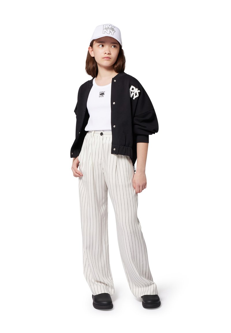 Chaqueta bomber negra, camiseta gráfica blanca, pantalones claros a rayas y zapatos negros, complementados por una gorra blanca con logo.