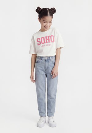 Dziewczyna z podwójnymi koczkami, ubrana w białą koszulkę z napisem "SOHO New York", jasnoniebieskie dżinsy i białe trampki, stoi i się uśmiecha.