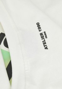 Camiseta blanca con el texto "ATELIER 1990 PARIS" en negro. Tejido de algodón suave con una textura lisa y un diseño gráfico colorido debajo.