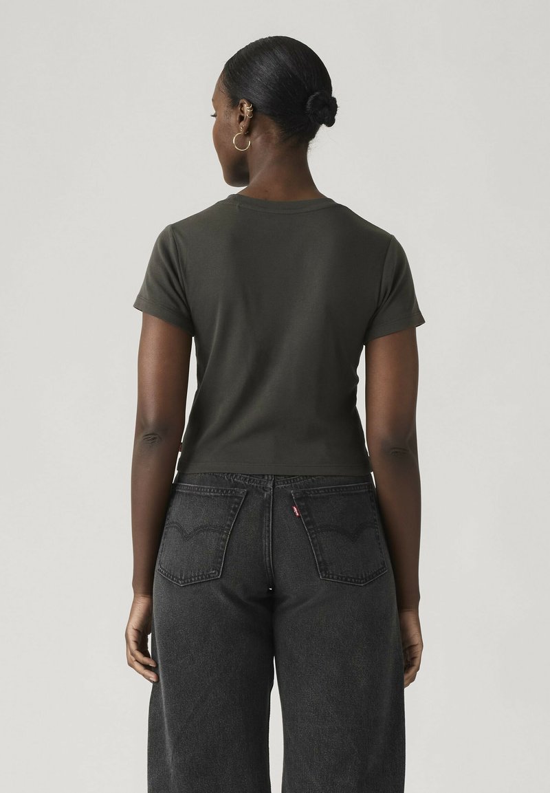 Vrouw met een donkere huid en haar in een knot draagt een donkergroen T-shirt met korte mouwen en hoog getailleerde zwarte spijkerbroek, kijkt weg van de camera.