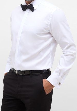 Hombre vistiendo una camisa blanca ajustada con pajarita negra, pantalones negros y cinturón negro, con las manos en los bolsillos, frente a un fondo liso.