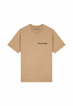 Jonge vrouw met steil blond haar, gekleed in een beige oversized T-shirt met de tekst 'YOU & ME TONIGHT' en blauwe spijkerbroek tegen een effen achtergrond.