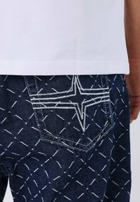 Primo piano di jeans blu scuro con un motivo a diamante cucito in bianco e un design a stella cucito sulla tasca posteriore, indossati con una camicia bianca.
