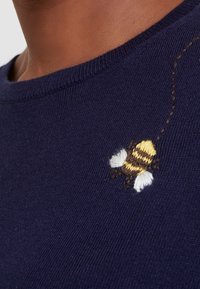 Marinblå tröja med en liten broderad bi i gult och vitt med svarta accenter, placerad nära halsringningen. Mjuk stickad textur.