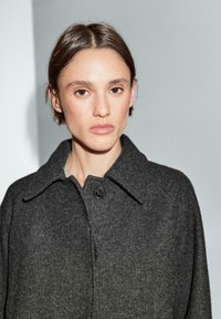 Szary wełniany płaszcz z kołnierzem, zapinany na jeden guzik, o teksturowanym wykończeniu. Tkanina wykazuje delikatny gradient od ciemnoszarego do jasnoszarego.