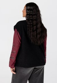 Zwarte mouwloze vest met een gestructureerd oppervlak, gedragen over een lange mouwen bordeauxrood shirt met geborduurde bladdesigns op de mouwen.