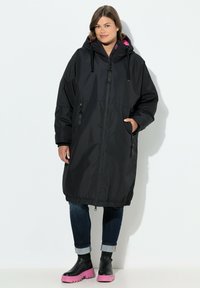 Abrigo negro impermeable con capucha, que presenta detalles en rosa, bolsillos con cremallera y un dobladillo elástico. Llevado sobre unos vaqueros y botas con suela rosa.
