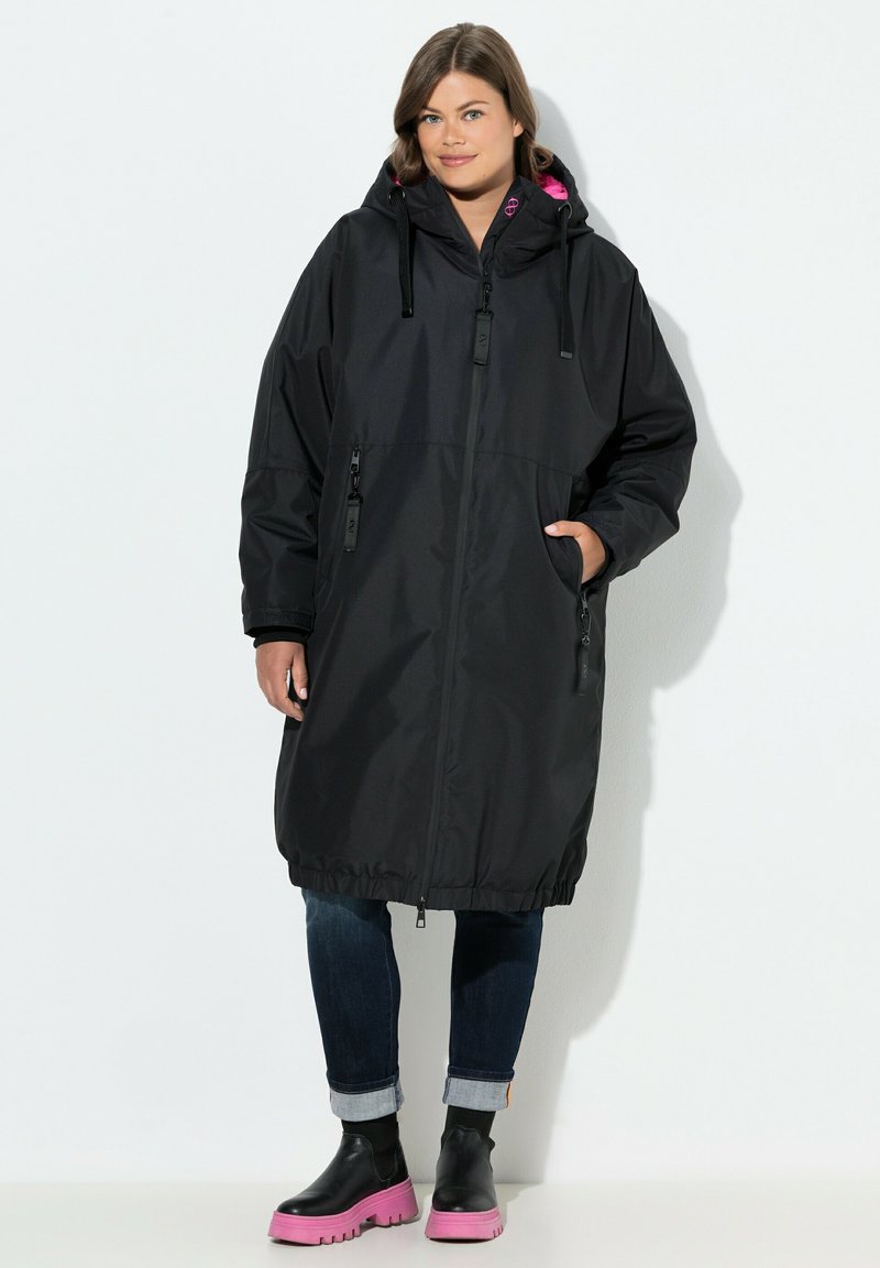 Abrigo negro impermeable con capucha, que presenta detalles en rosa, bolsillos con cremallera y un dobladillo elástico. Llevado sobre unos vaqueros y botas con suela rosa.