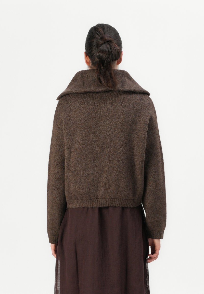 Bruine, gestructureerde gebreide trui met een brede kraag en een cropped zoom. Heeft lange mouwen en een ontspannen pasvorm, gecombineerd met een donkere jurk.