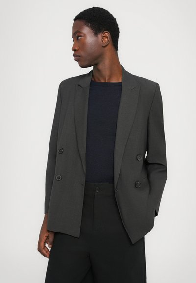 Γκρι διπλόστιχο σακάκι με δύο μαύρα κουμπιά, κομψός σχεδιασμός, μακριά μανίκια και πέτο, φορεμένο πάνω από ένα σκούρο μπλε πλεκτό τοπ.