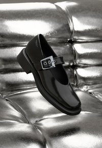 Black leather Mary Jane shoe with a round toe, adjustable silver buckle strap, and flat rubber sole, set against a metallic background.