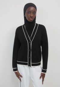 Frau, die eine schwarze Knopfleiste mit weißen Besätzen, einen schwarzen Hijab und weiße Hosen trägt, steht vor einem schlichten hellen Hintergrund.