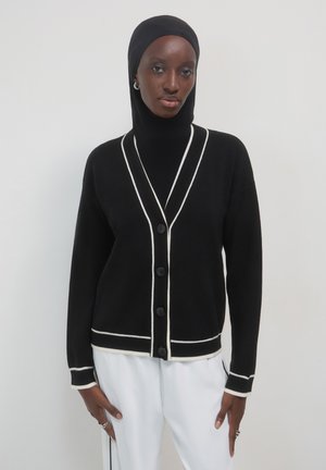 Frau, die eine schwarze Knopfleiste mit weißen Besätzen, einen schwarzen Hijab und weiße Hosen trägt, steht vor einem schlichten hellen Hintergrund.