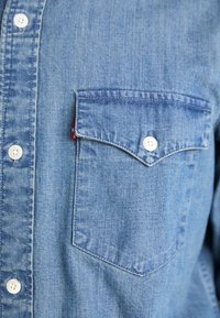 Camicia di jeans azzurro chiaro con bottoni bianchi e una tasca sul petto con bottoni, caratterizzata da un'etichetta rossa sul lato sinistro.