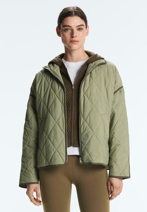 Frau trägt eine hellgrüne gesteppte Jacke über einem braunen Reißverschluss-Pullover und beigen Leggings, steht vor einem schlichten, hellen Hintergrund.