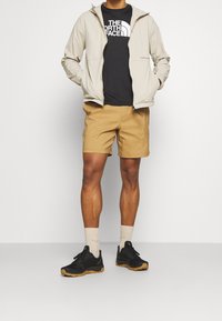 Světle béžová kapuce bunda, černé tričko s grafikou, béžové šortky a černé běžecké boty s gumovou podrážkou. Ponožky jsou světle béžové.