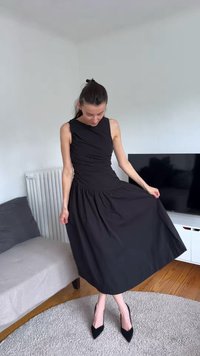 Naine seljas musta varrukateta midi kleidi ja mustade teravate kontsadega seisab elutoas ümmargusel vaibal.