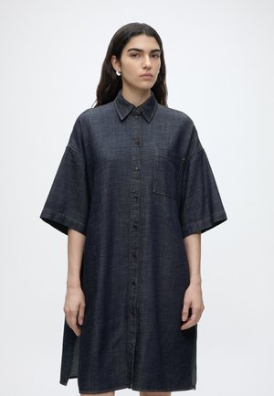Frau mit langen dunklen Haaren, die ein lockeres, dunkles Denim-Kleid mit Knöpfen, kurzen Ärmeln und einer Brusttasche trägt, stehend vor einem schlichten weißen Hintergrund.