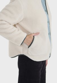 Chaqueta de forro polar en crema con un frente acolchado azul y ribete gris. Presenta un bolsillo de parche y un acabado suave y texturizado.