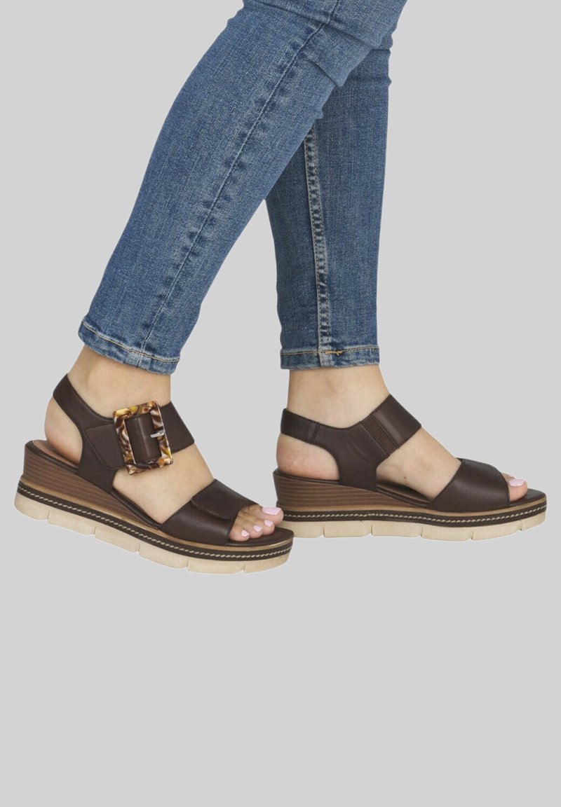 Pieds portant des sandales compensées marron avec une grande boucle et une semelle claire, associées à un jean skinny bleu sur un fond uni.