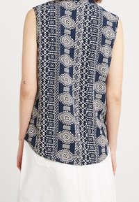 Blouse sans manches bleu marine avec un motif géométrique et floral blanc, tissu léger et ourlet incurvé.